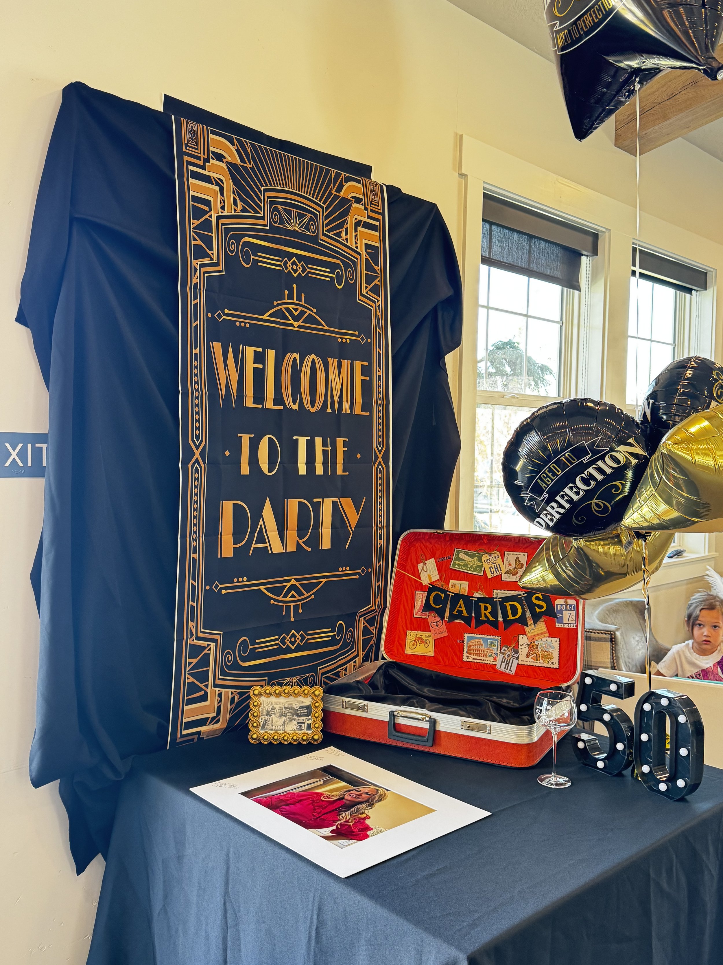 Decorative sign reading "Welcome to the Party" on a wall, with balloons and a table displaying various items including a red vintage-style suitcase, photocopies, a framed photo, and a photo album, near windows with dark blinds.