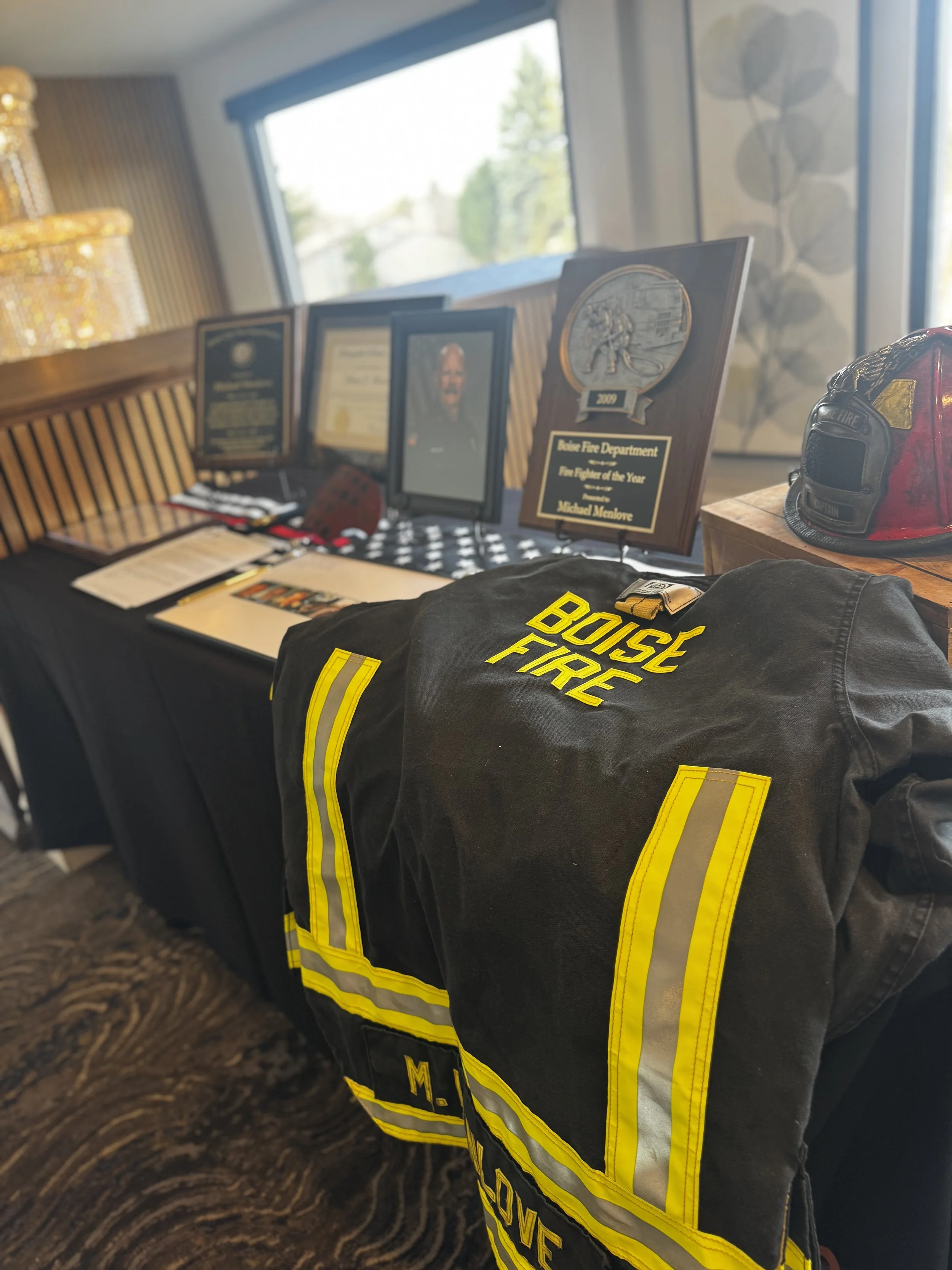 Display table with firefighter memorabilia including a plaque, framed certificate and photo, firefighter helmet, a firefighter jacket, and a hat, in a well-lit room with large windows.