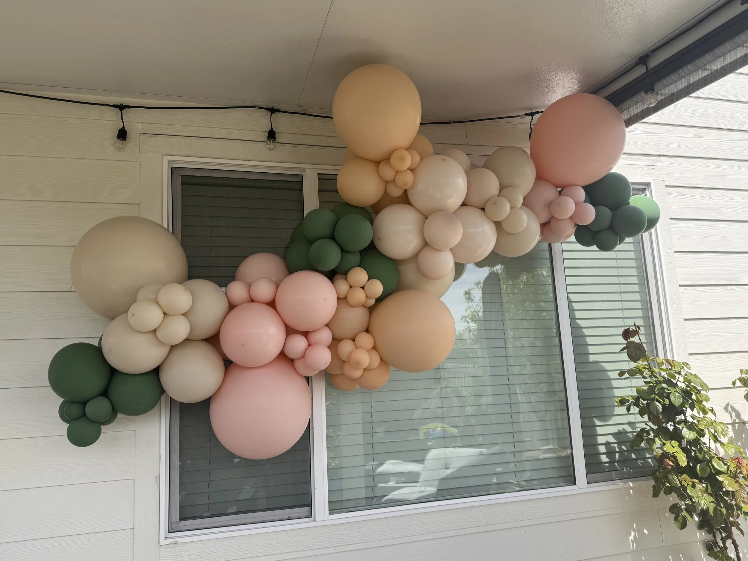Decorative balloon arrangement with various sizes and pastel colors, attached above a sliding glass door on an exterior wall of a house with white siding and a window.