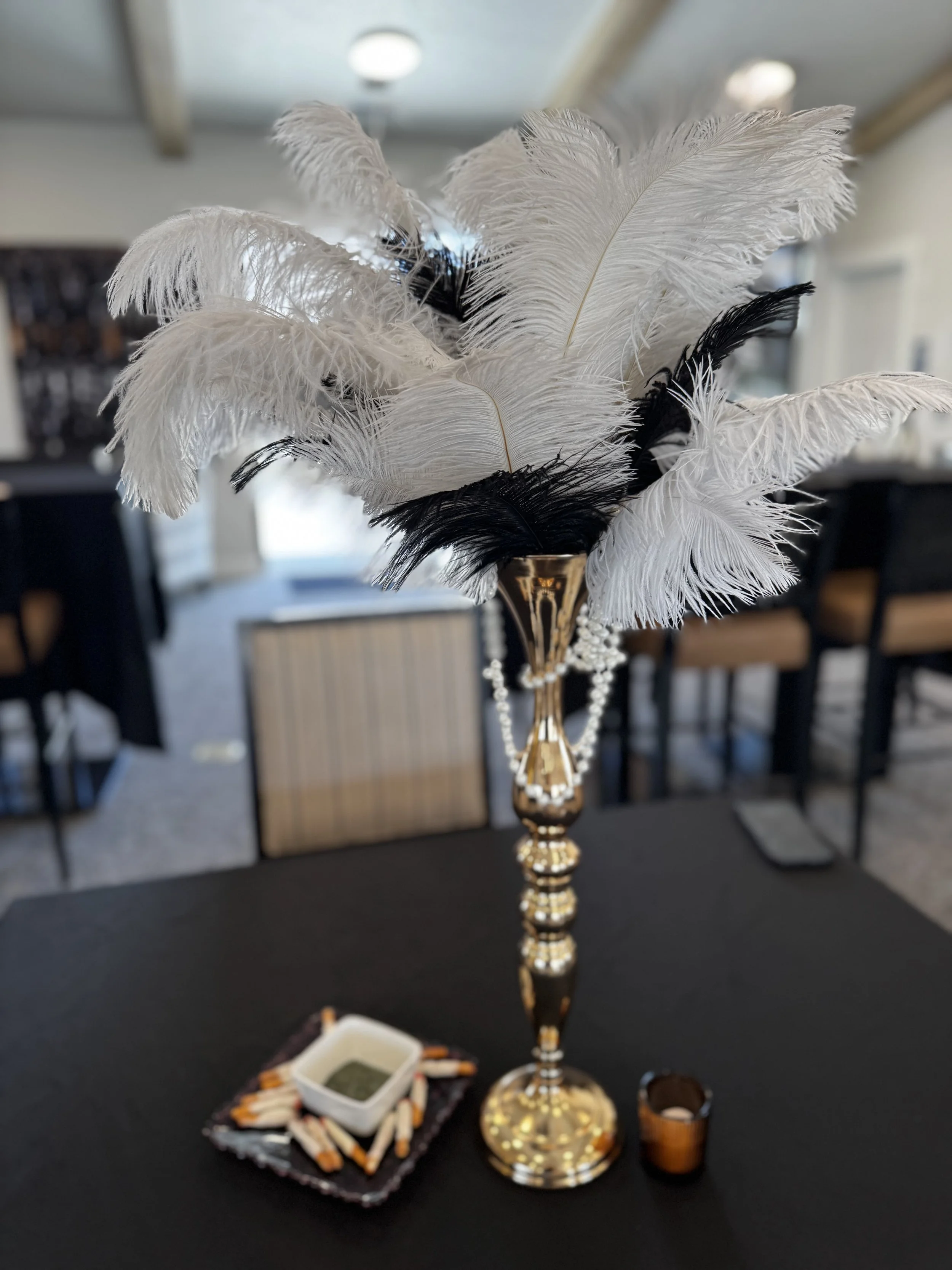 A close-up of a decorative vase filled with white and black feathers, placed on a black table. In front of the vase, there are small containers with various items, and a beaded jewelry piece. The background shows a room with chairs and tables.
