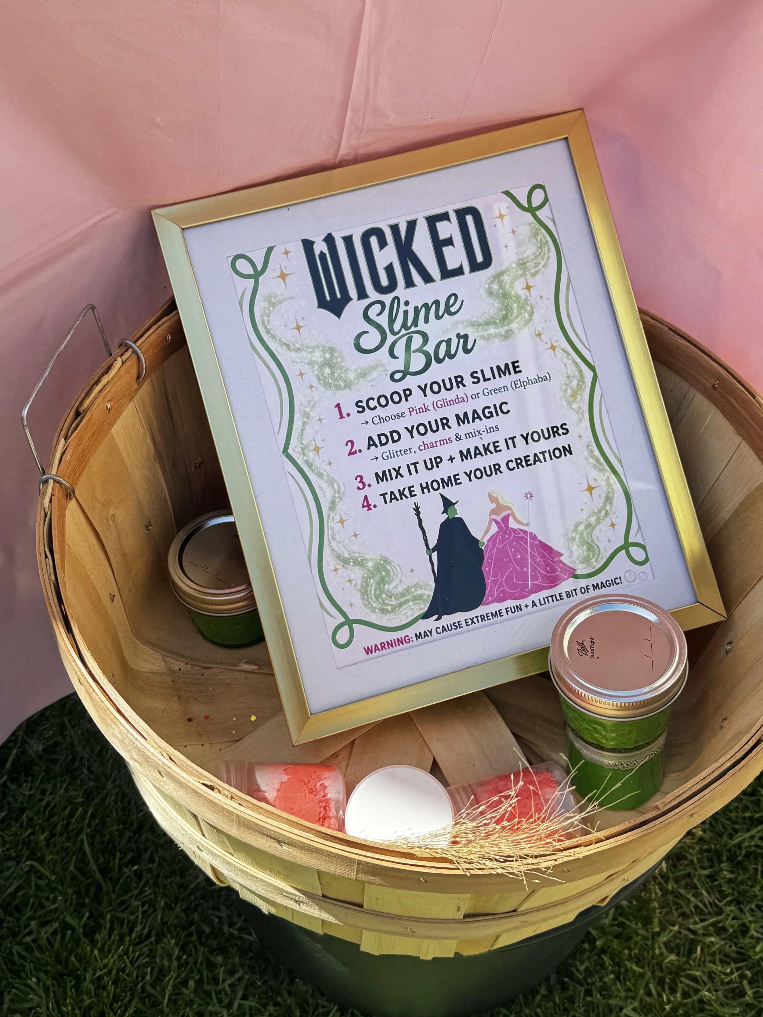 A wooden bucket with a sign for a 'Wicked Slime Bar' inside. The sign provides instructions for making slime, including scooping slime, adding glitter and charms, mixing, and taking it home. Several small containers of slime are placed inside the buc