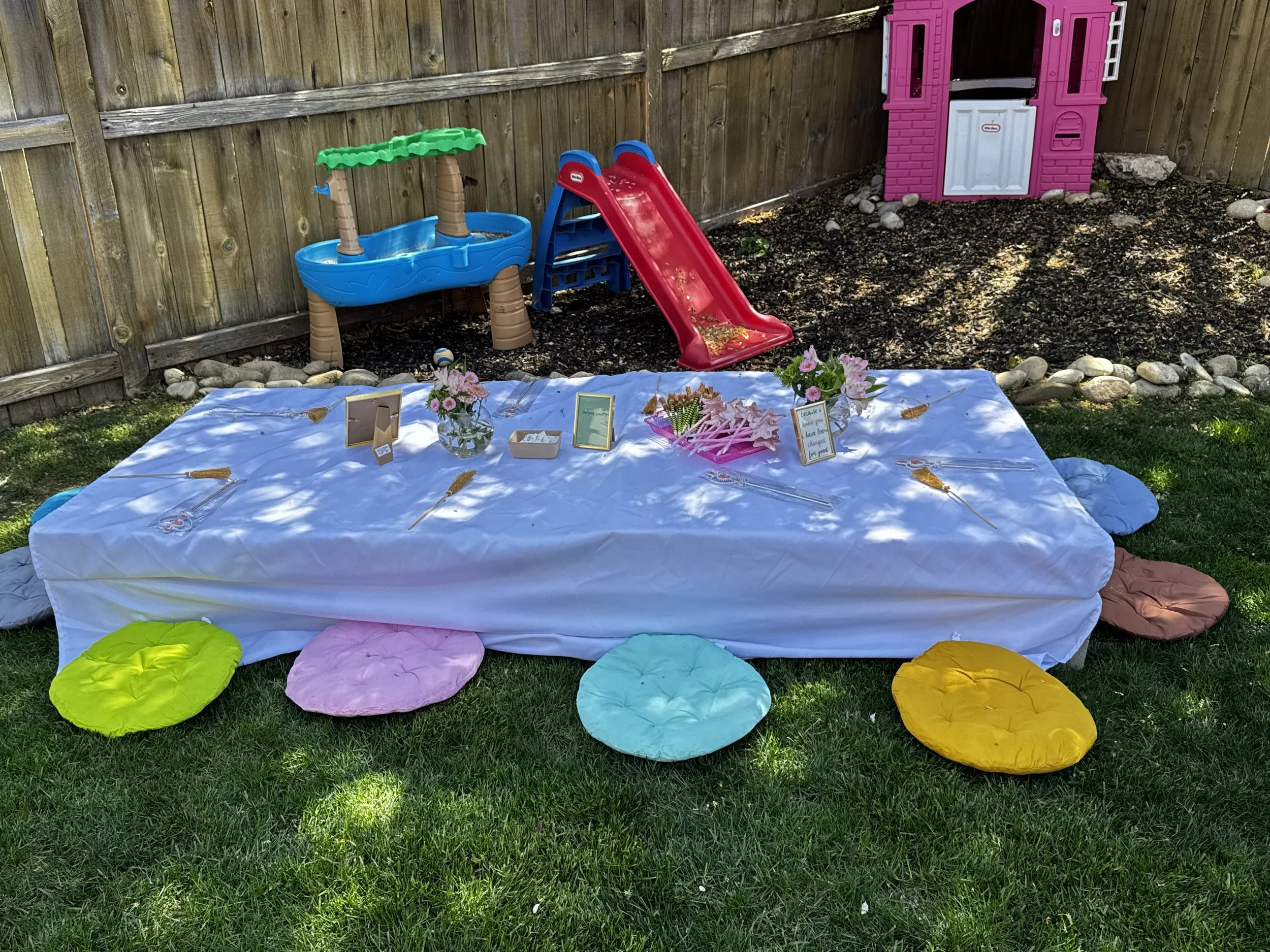 A backyard with a white blanket on the grass, colorful round cushions, a small pink and gray playhouse, some flowers, and a table set for a celebration or gathering.