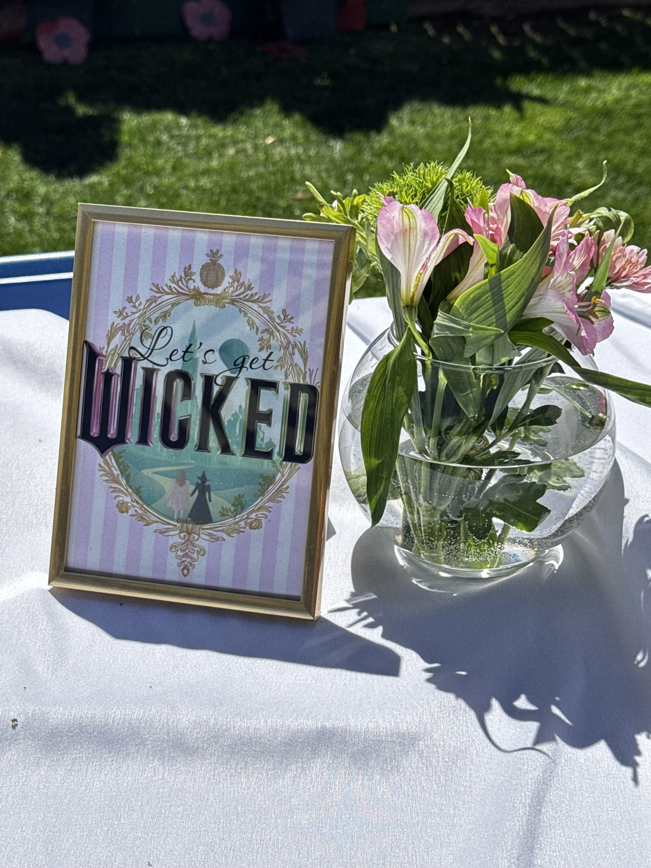 A table with a framed sign that says 'Let's get WICKED' and a flower arrangement in a glass vase, outdoors on a sunny day.
