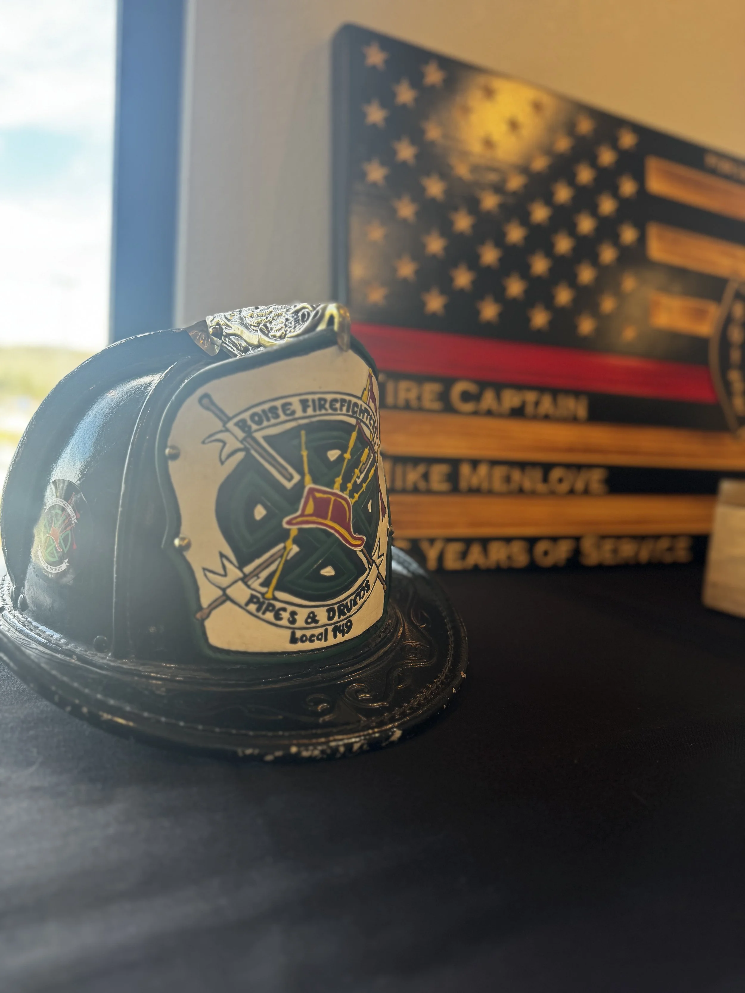 A firefighter's badge features a snake wrapped around a sword, with the words 'Boise Firefighters Pipes & Drums Local 119.' In the background, there is a wooden plaque with an American flag design and the words 'Fire Captain Mike McLove 50 Years of S