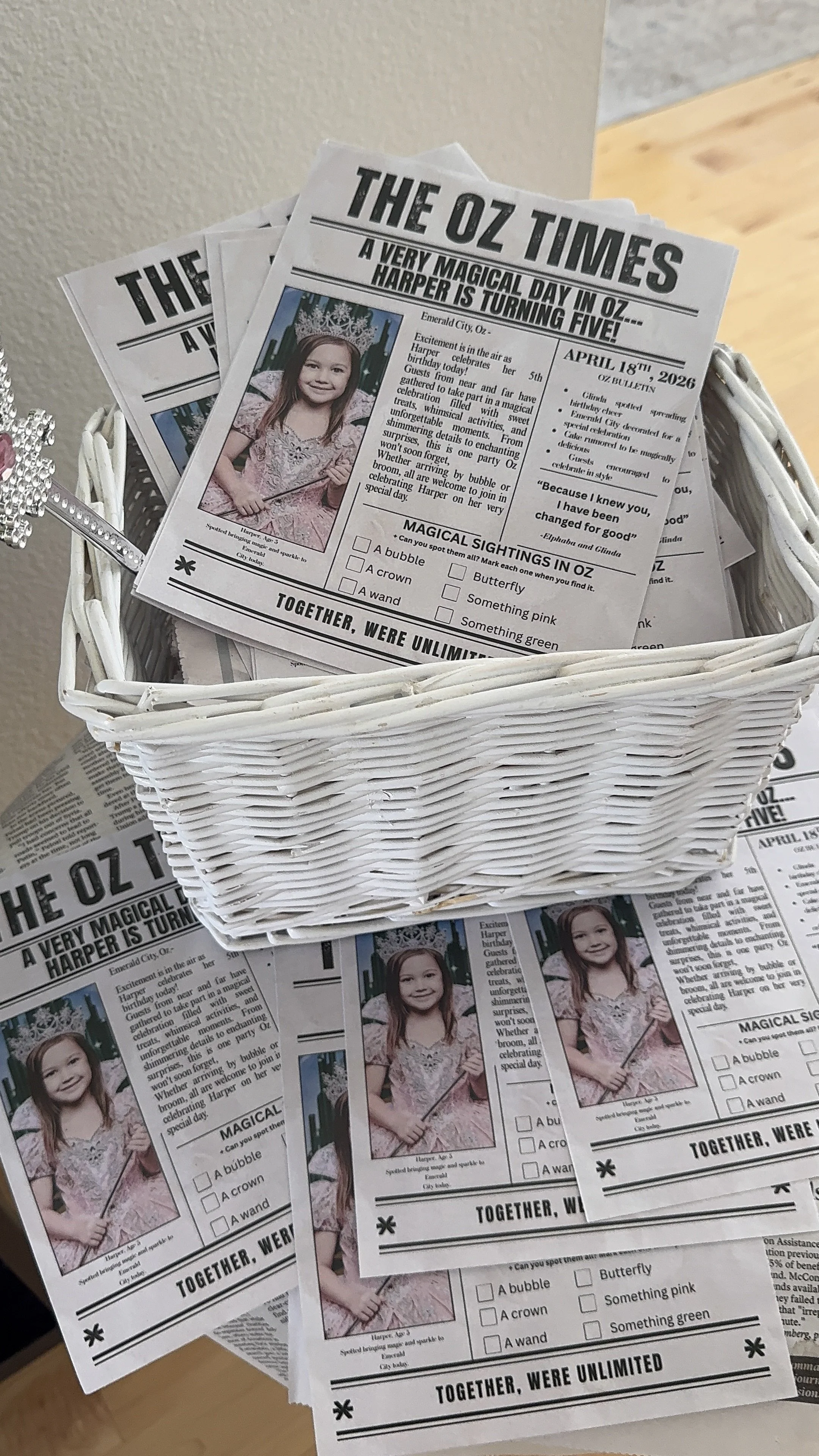 A stack of newspapers titled 'The Oz Times' with a photo of a young girl wearing a crown and holding a wand, placed in a white basket on a wooden table.
