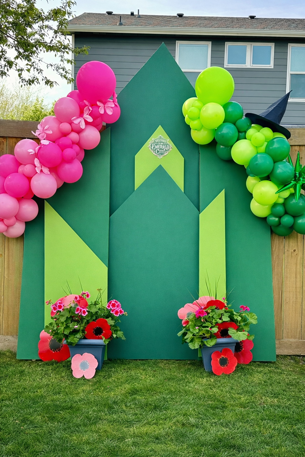 Green castle-themed backdrop decorated with pink and green balloons, complemented by potted pink and red flowers in front, and a wizard's hat on the right.
