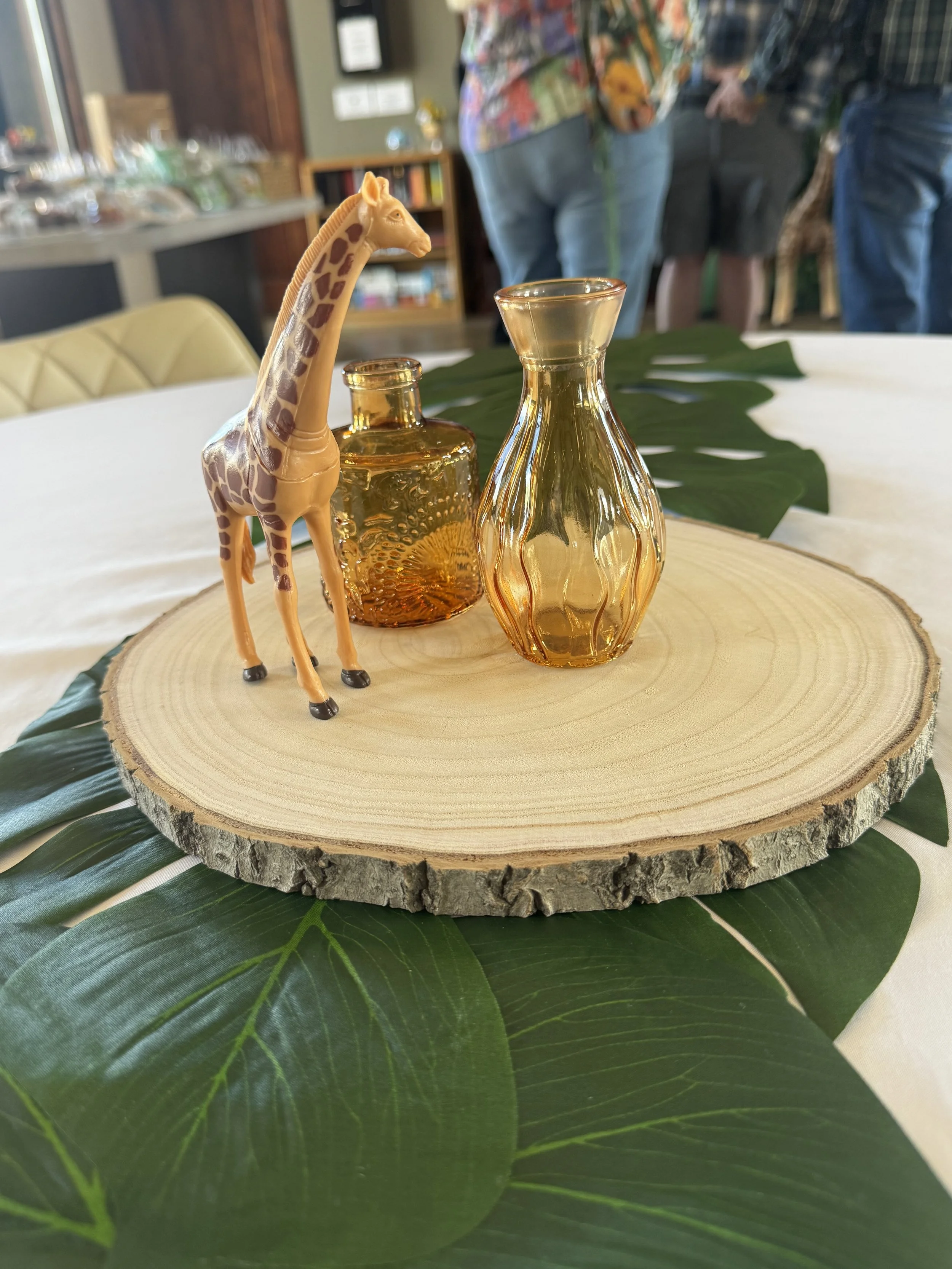 Decorative giraffe figurine on a wooden round slab with glass vases, green leaves, and a blurred indoor background.