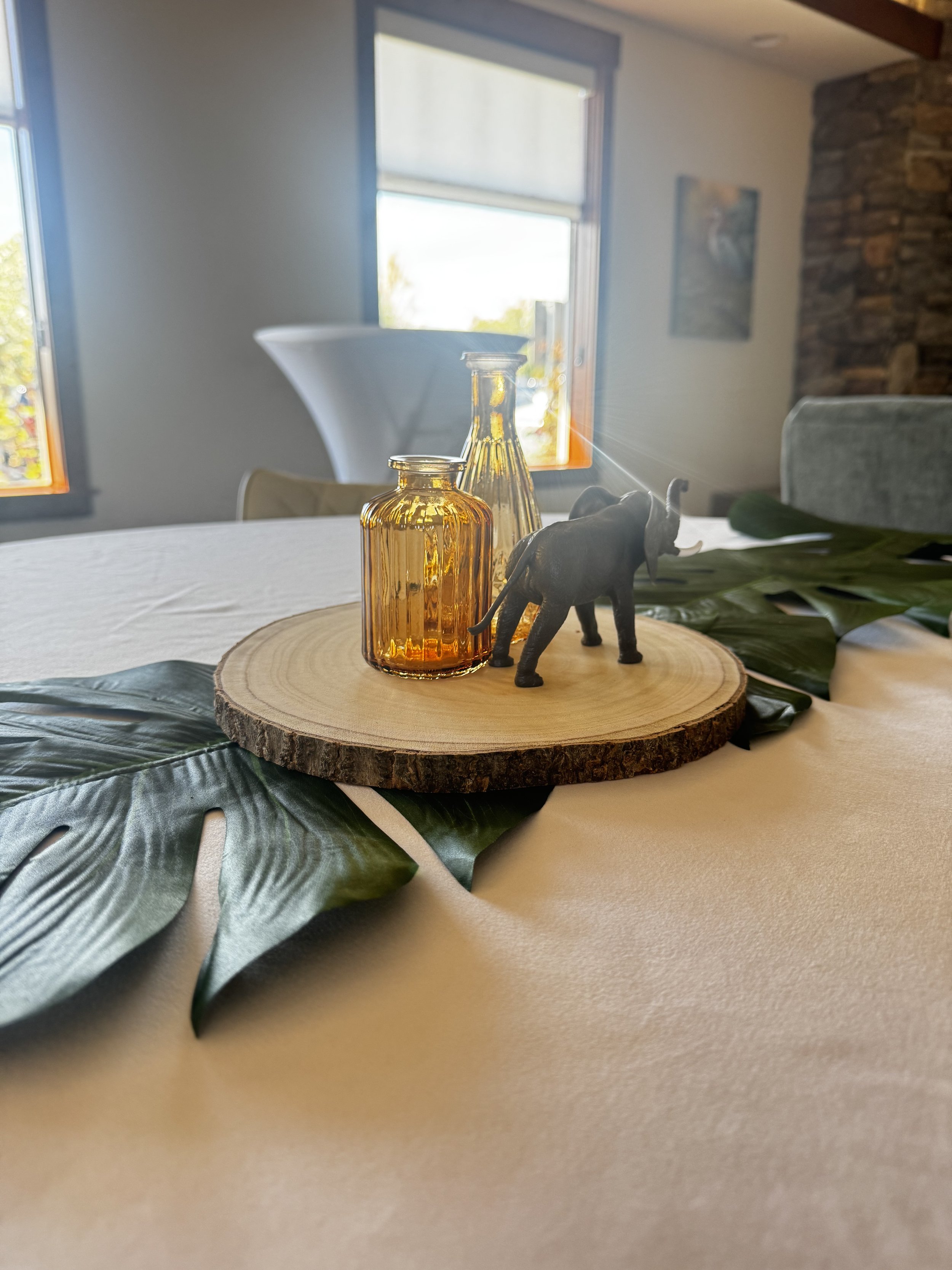 Decorative bottles and elephant figurine on a wooden circular base, placed on a table with large green leaves, in a room with windows and a stone wall.
