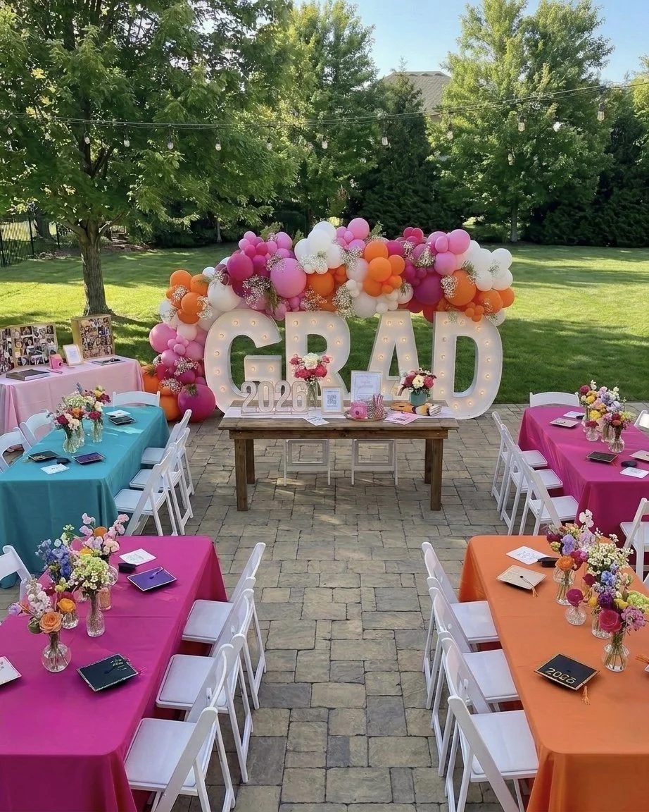 Backyard graduation party setup with colorful tables and a balloon backdrop spelling 'GRAD' and '2026'.