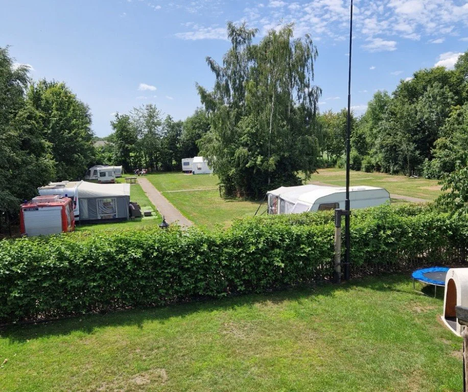 Een camping met caravans en campers op een groene, zonnige dag, omgeven door bomen en een heg.