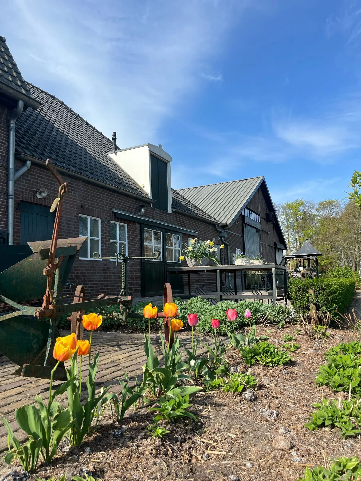 Bloemen in een tuin voor een boerderij met een donkere bak en een brug onder een blauwe hemel.