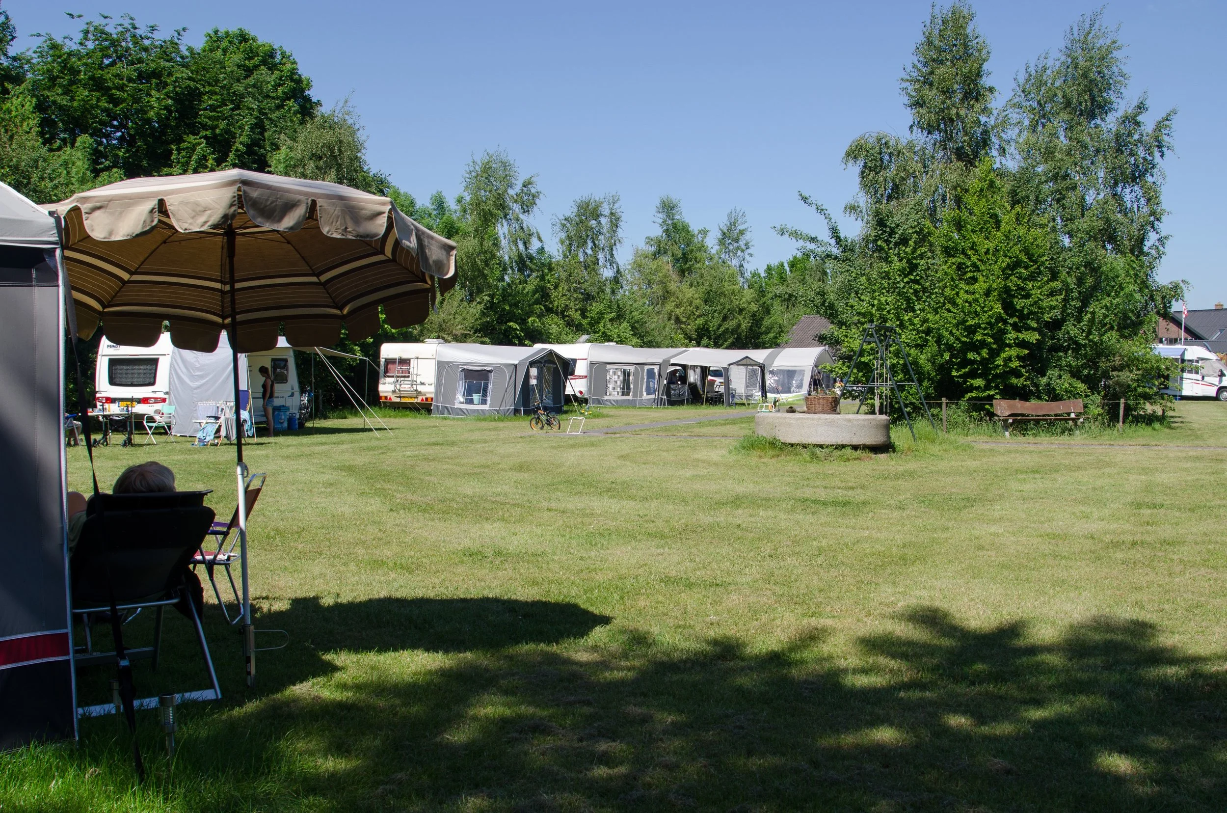 Een camping met caravans en tenten, groene bomen en een grasveld, onder een blauwe hemel, met een schommel en een bank.