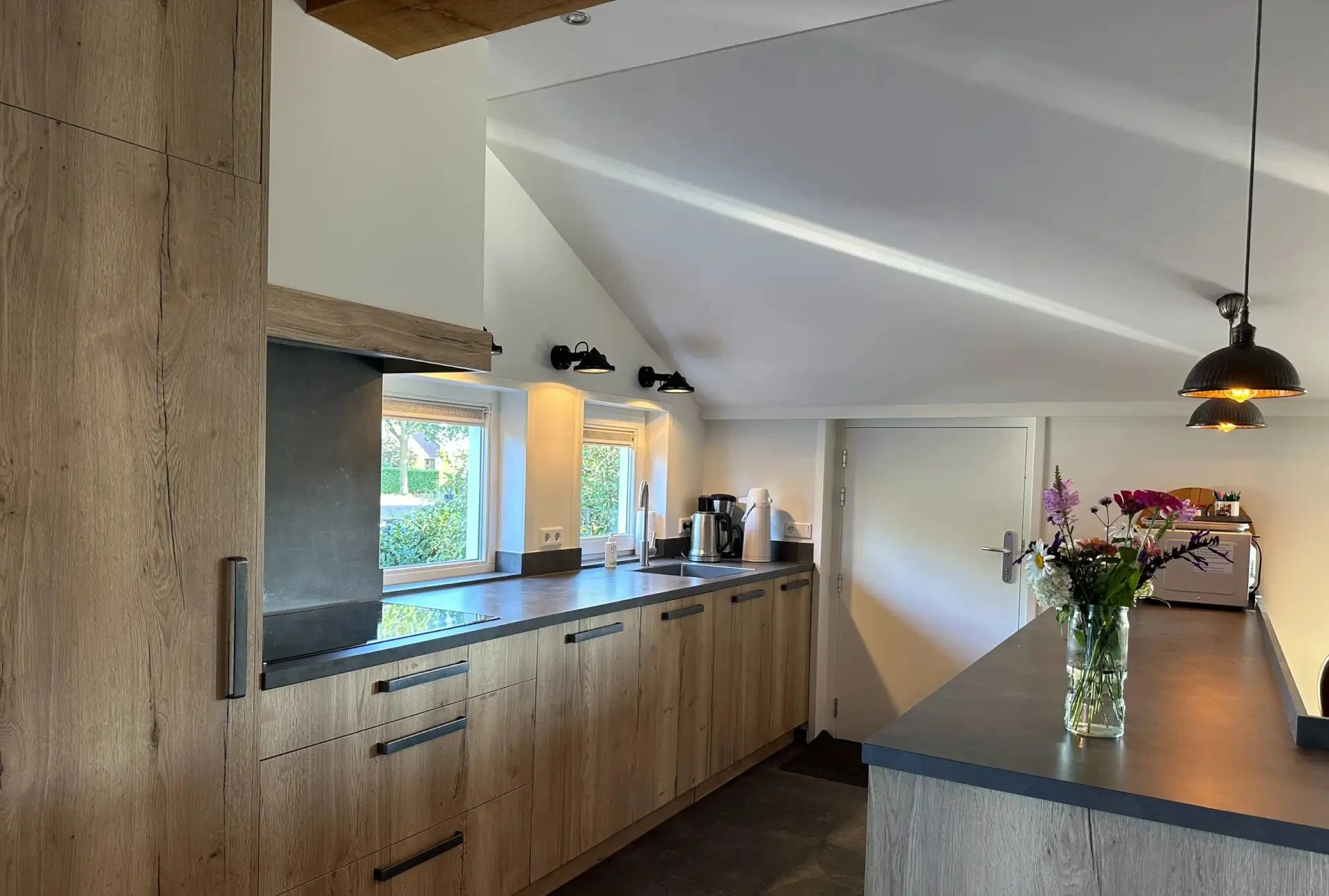 Een moderne keuken met houten kasten, een werkblad, twee ramen, een bloemenvaas, en apparaten zoals een koffiezetapparaat en een magnetron.