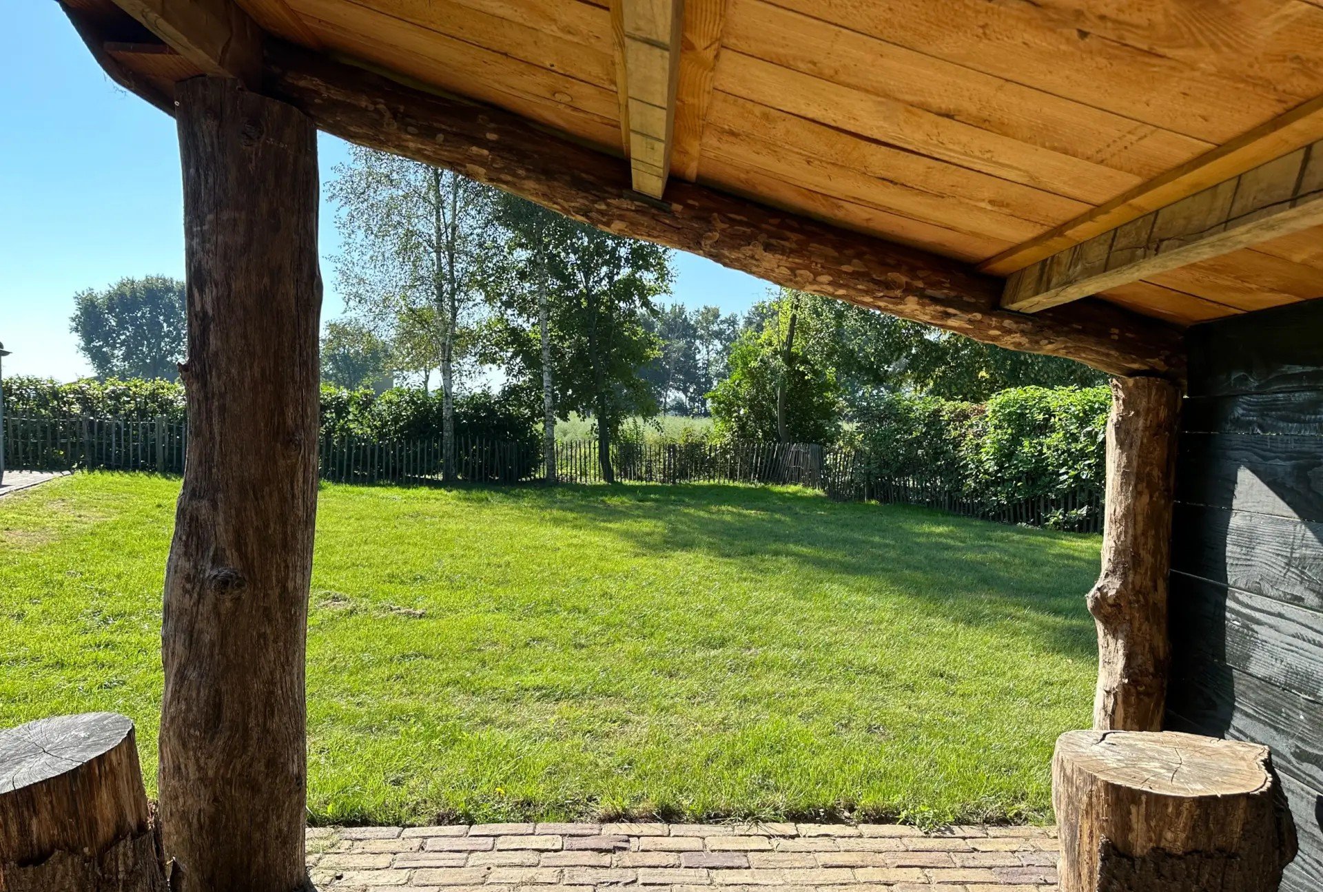 Een tuin met een groene plaggen, omgeven door een houten schutting en diverse bomen, gezien vanaf onder een houten overkapping met grote houten palen.