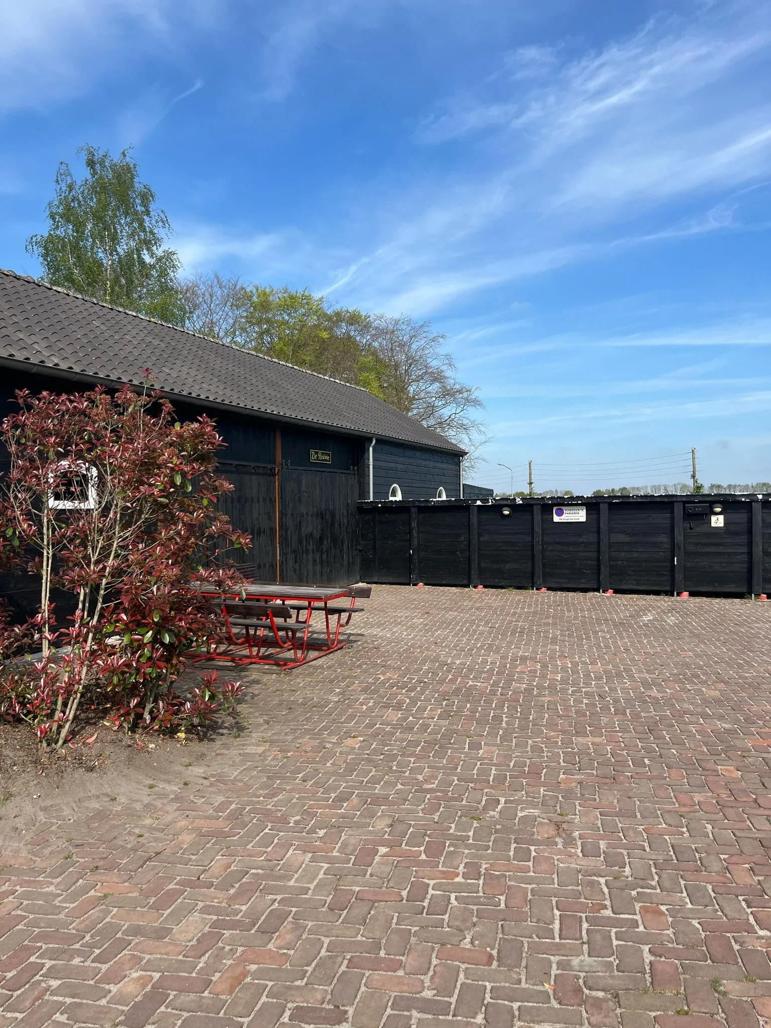 Een open binnenplaats met een betonnen tegelvloer, een zwarte houten schuur, een houten picknicktafel met bankjes, een rode struik links, een blauwe lucht met witte wolken, en bomen op de achtergrond.