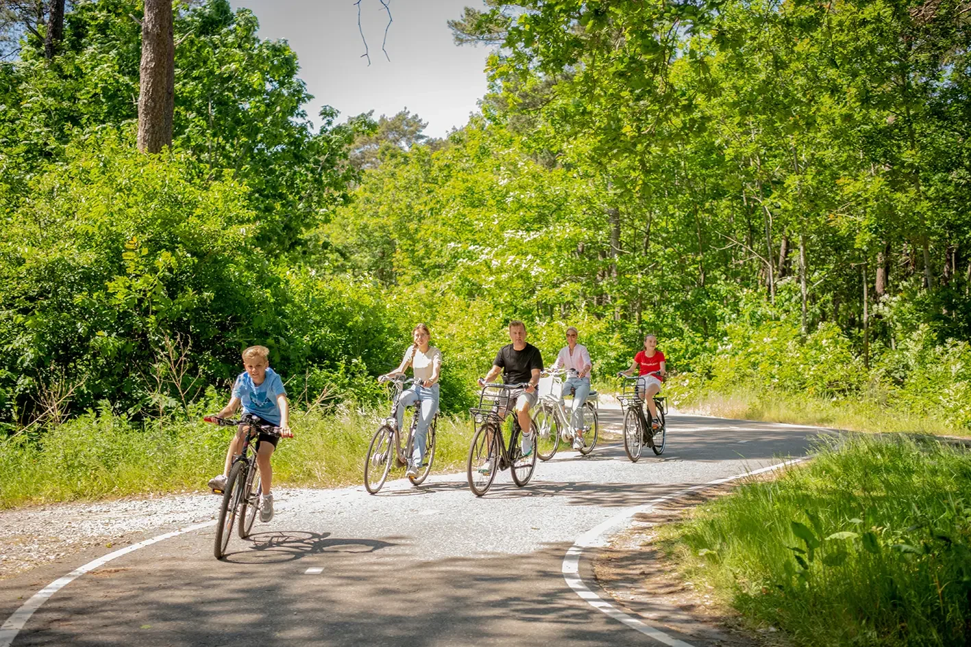 Vijf mensen fietsen samen op een bosweg, omringd door groene bomen en struiken, op een heldere dag.
