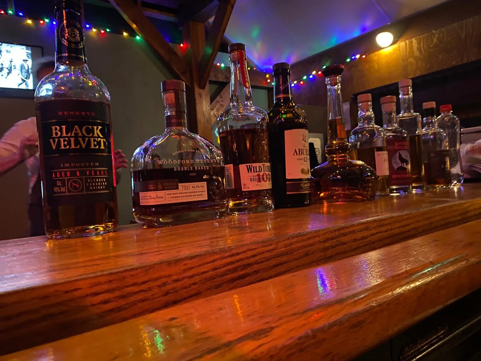 Verschillende whisky flesjes op een houten bar, decoratief verlicht met kleurrijke feestverlichting.