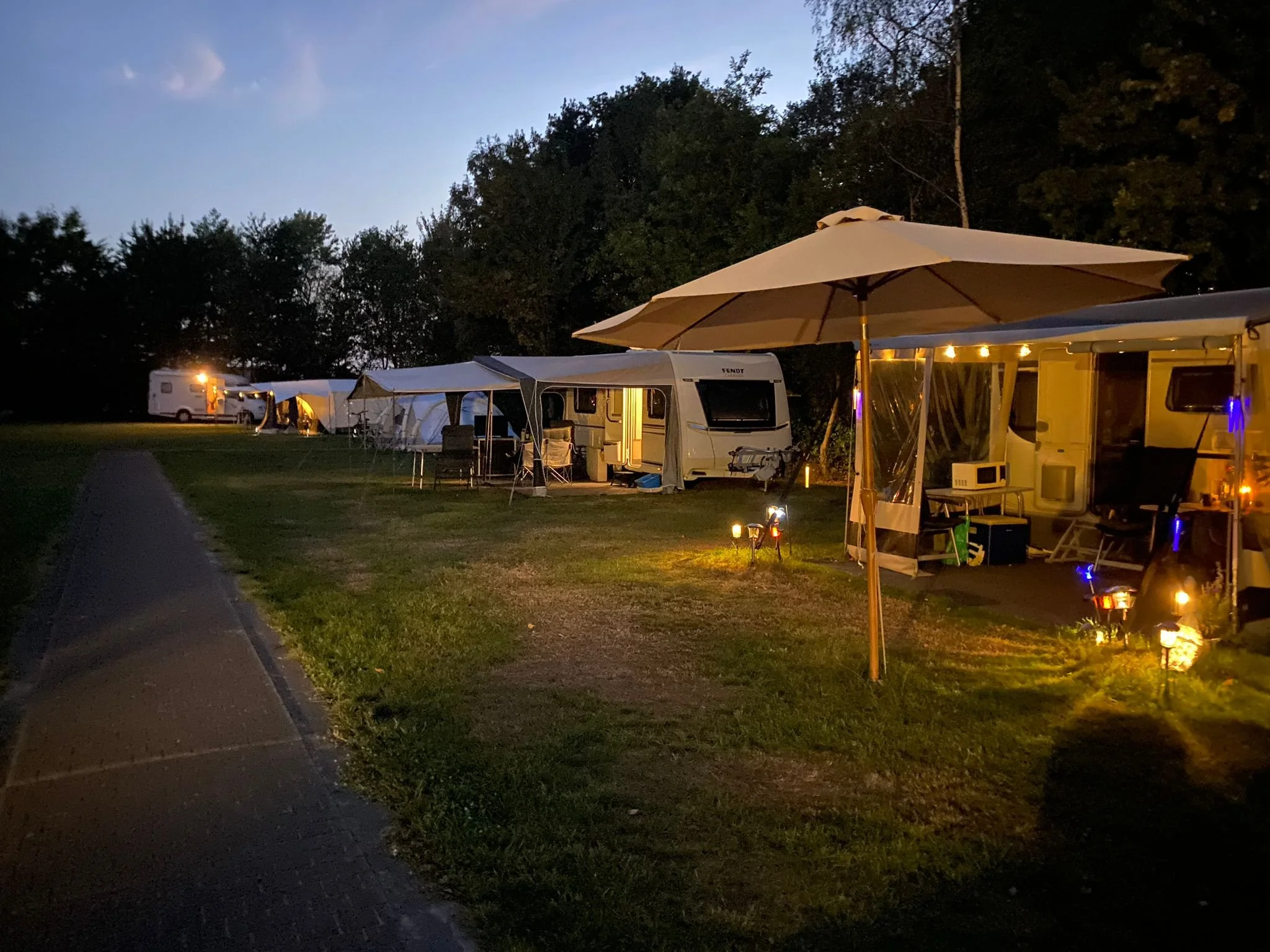 Een camping bij avond, met caravans, tenten en een parasol die verlicht zijn door kleine lampjes.