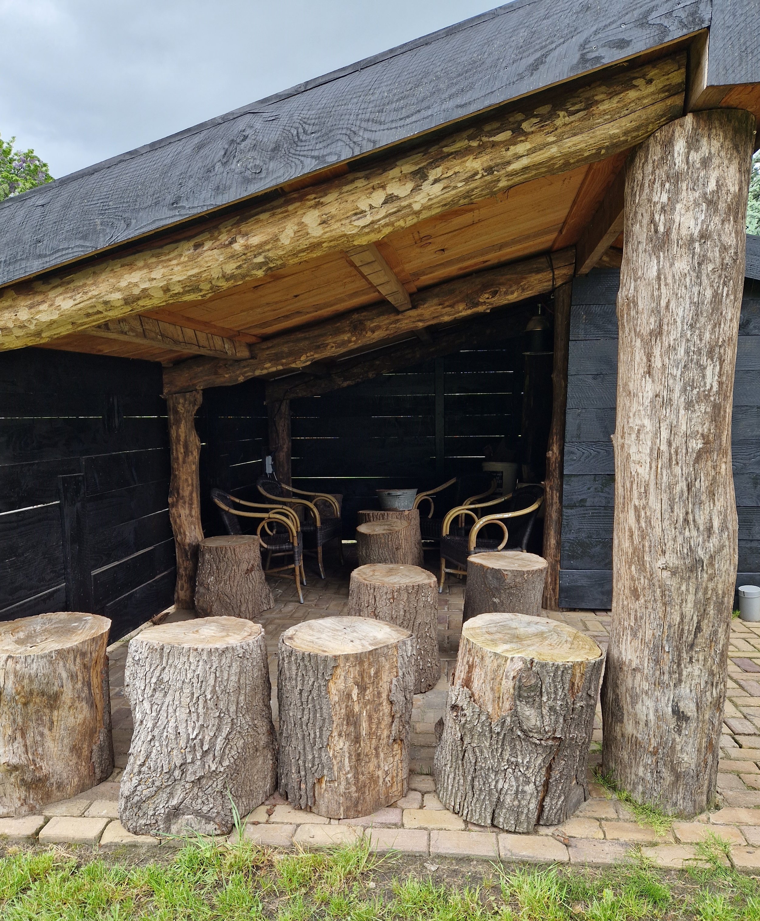 Een houten veranda met buitenzitplek, bestaande uit een kap met een houten dak en stammen als steunpilaren. Op de grond staan boomstammen als zitplaatsen en er are een paar stoelen op de achtergrond.