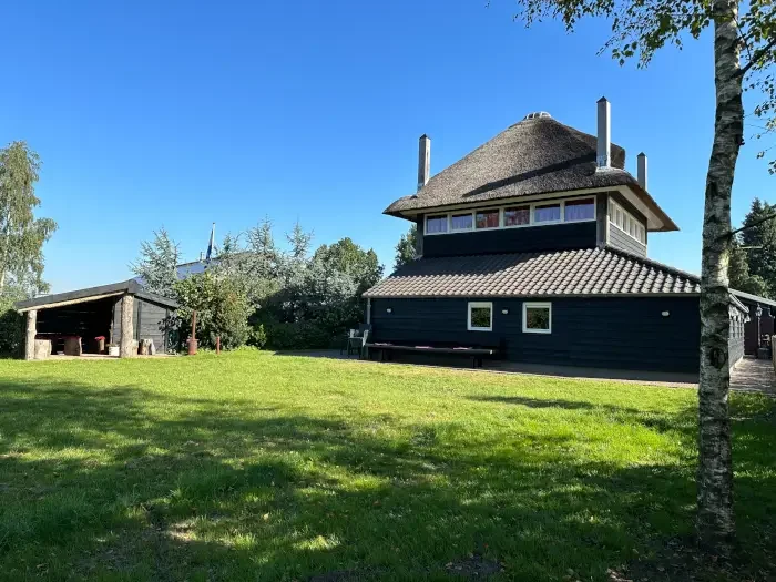 Een traditionele Nederlandse woning met een rieten dak en een grote groene tuin onder een blauwe hemel.