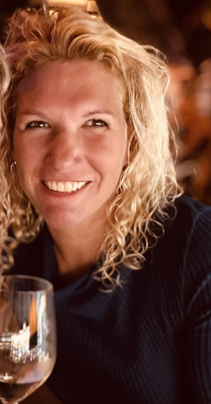 Close-up van een lachende vrouw met blonde, krullende haar, die een glas rode wijn vasthoudt, in een gezellige binnenruimte.