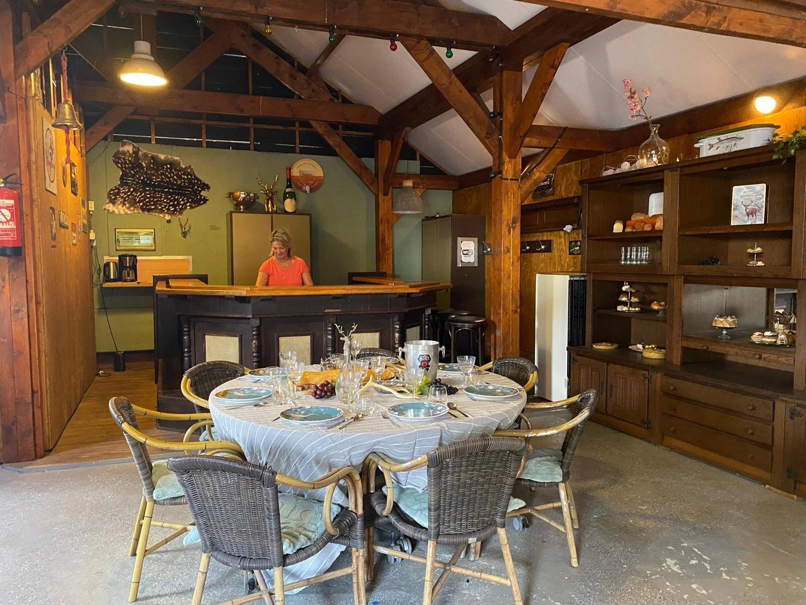 Een eetkamer met een ronde tafel bedekt met een tafelkleed, klaargemaakt voor een maaltijd. De tafel heeft borden, glazen en bestek. Er zitten zes rieten stoelen om de tafel. In de achtergrond bevindt zich een bar met een vrouw achter de bar, en een 
