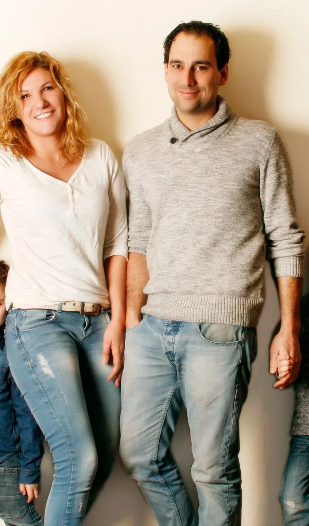 Twee mensen, een vrouw met rood, krullend haar in een witte blouse en een man met zwart, stekelig haar in een grijze trui, staan naast elkaar tegen een lichte wand, lachend en ontspannen.