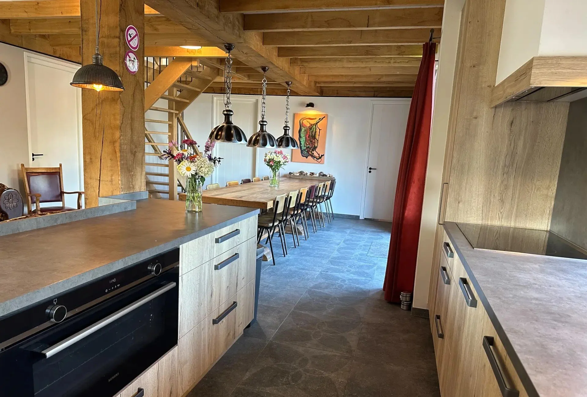 Een open keuken en eetkamer met houten meubels, bloemen en een schilderij aan de muur.