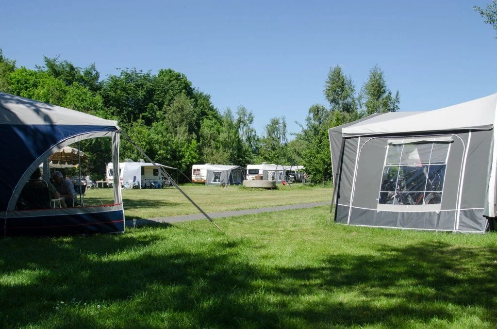 Tentencamping op een grasveld met meerdere caravans en tenten onder een blauwe hemel, omringd door groene bomen.