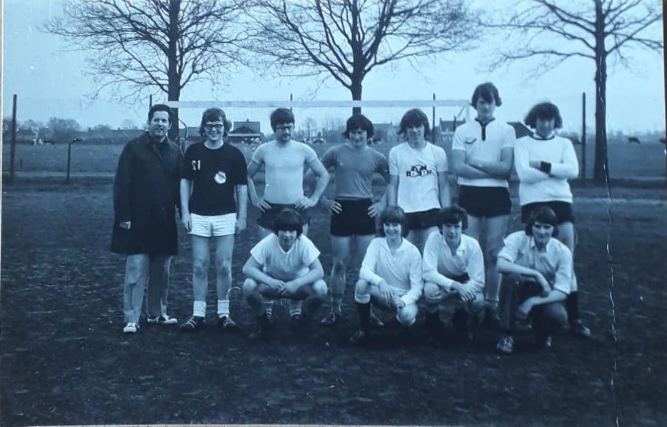 Tien kinderen en één volwassene op een sportschoolveld met volleybalnet, in een park met bomen op de achtergrond.