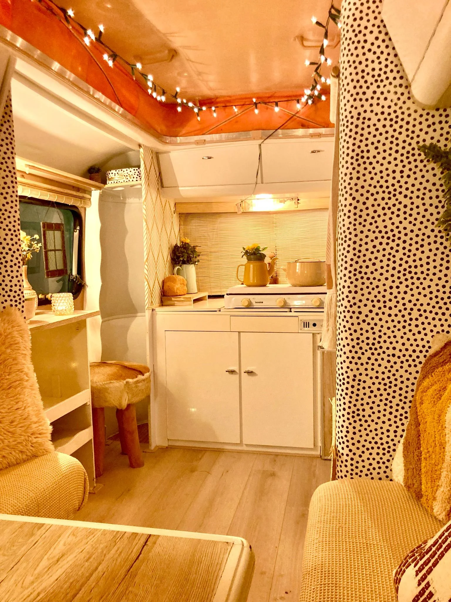 Gezellig ingerichte keuken met witte kasten, een geel gietijzeren kan, pannen, gekleurde stoelen met zachte kussens, een tafel, een raam met gordijn, en plafondverlichting en kerstverlichting.