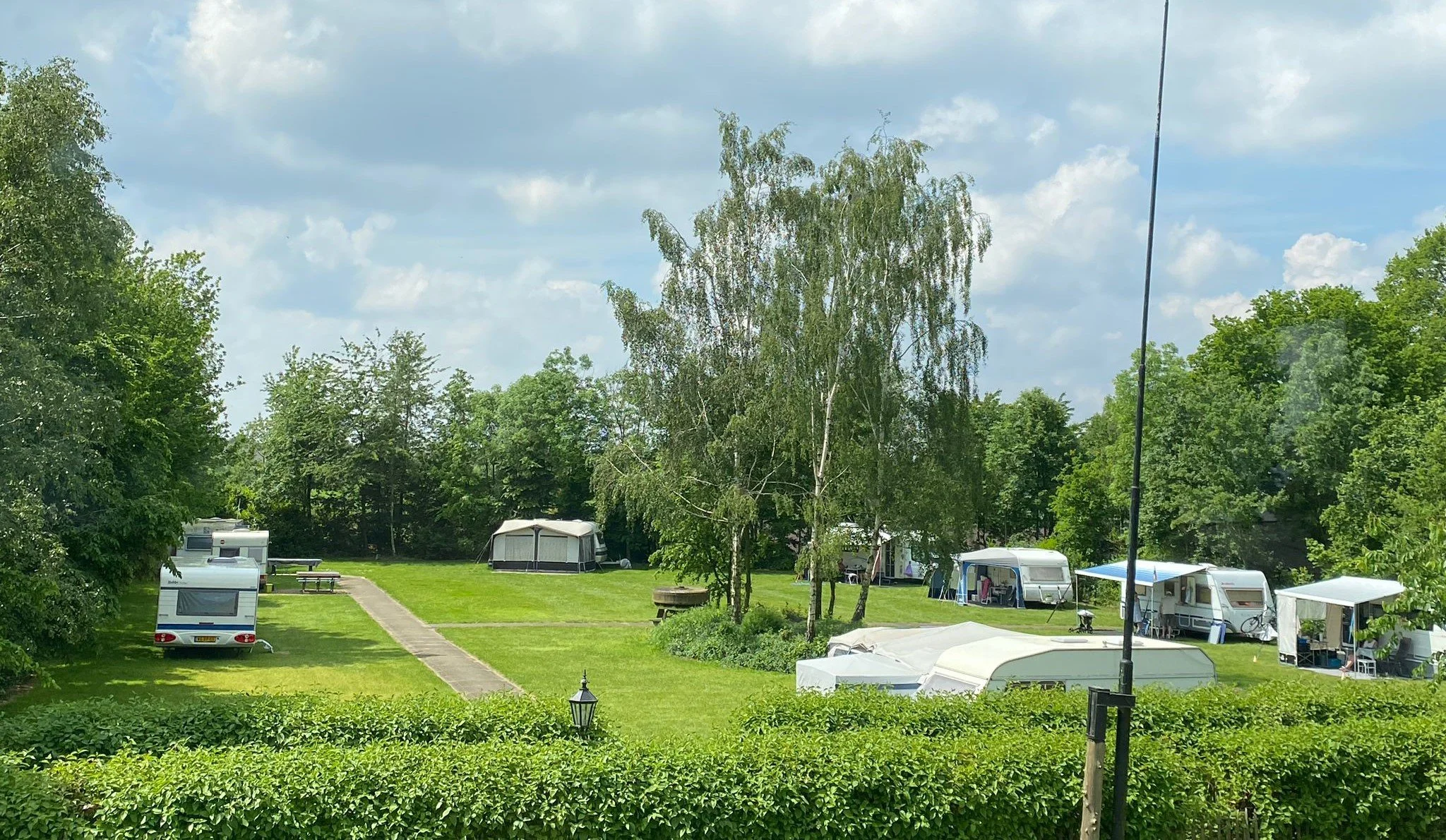 Een camping met caravans en tenten, omgeven door groene bomen en een grasveld, onder een bewolkte hemel.