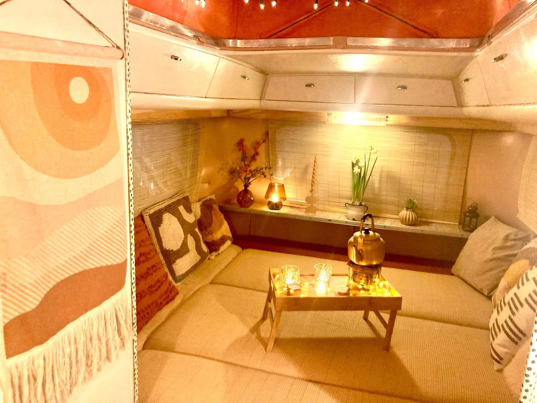 Knus woonkamer met lage bank, kussens, houten salontafel met kaarsen, thee-urn en decoratie, verlichting, planten en kussens met patronen.