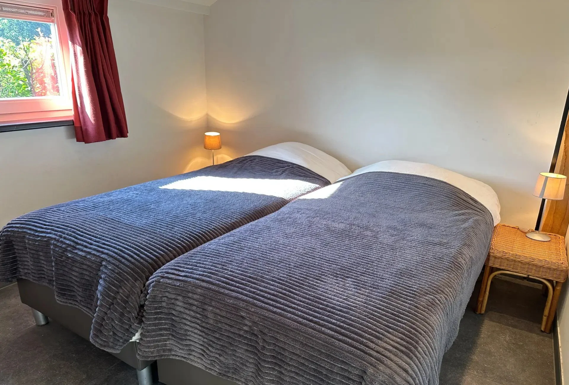 Twee bedden met een grijze deken, apart naast een raam met roze gordijnen, en een nachtkastje met een lamp.