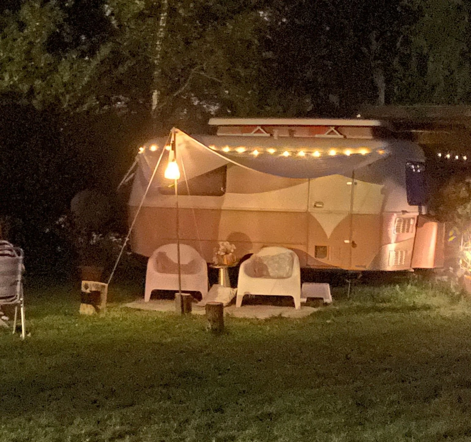 Een camper met een lichte binnenverlichting, onder een luifel met feestverlichting, in een tuin met gras. Er staan twee witte stoelen en een kleine tafel met bloemen erop.