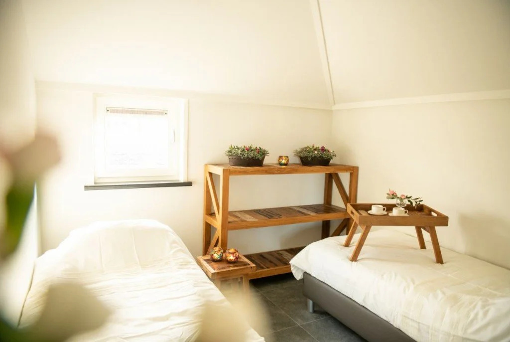 Twee bedden met witte beddengoed in een lichte kamer, met een houten rek en een dienblad met kopjes en bloemen.