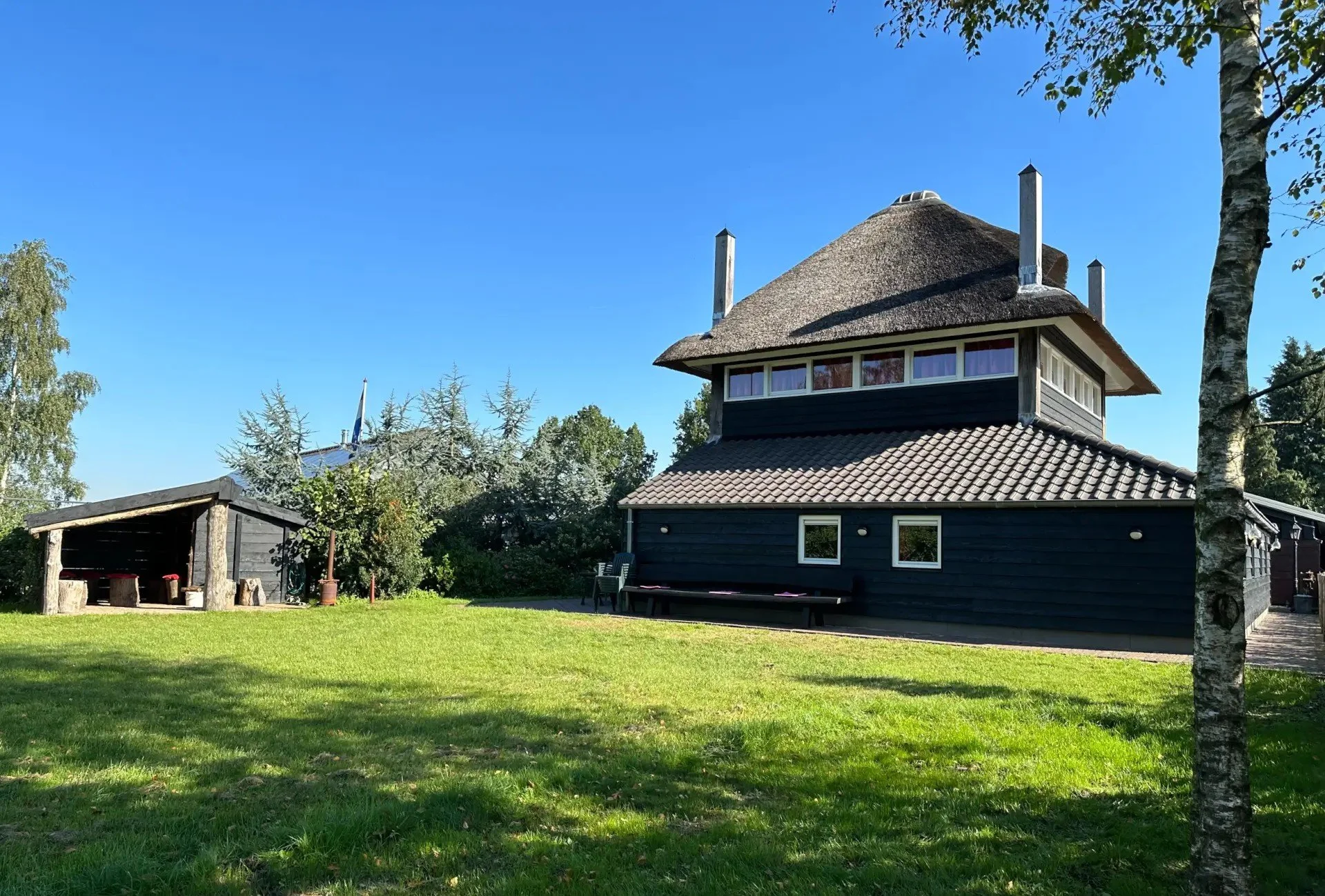 Een zwart houten huis met een thatchedak en veel ramen, omgeven door een groene tuin en bomen, onder een heldere blauwe hemel.