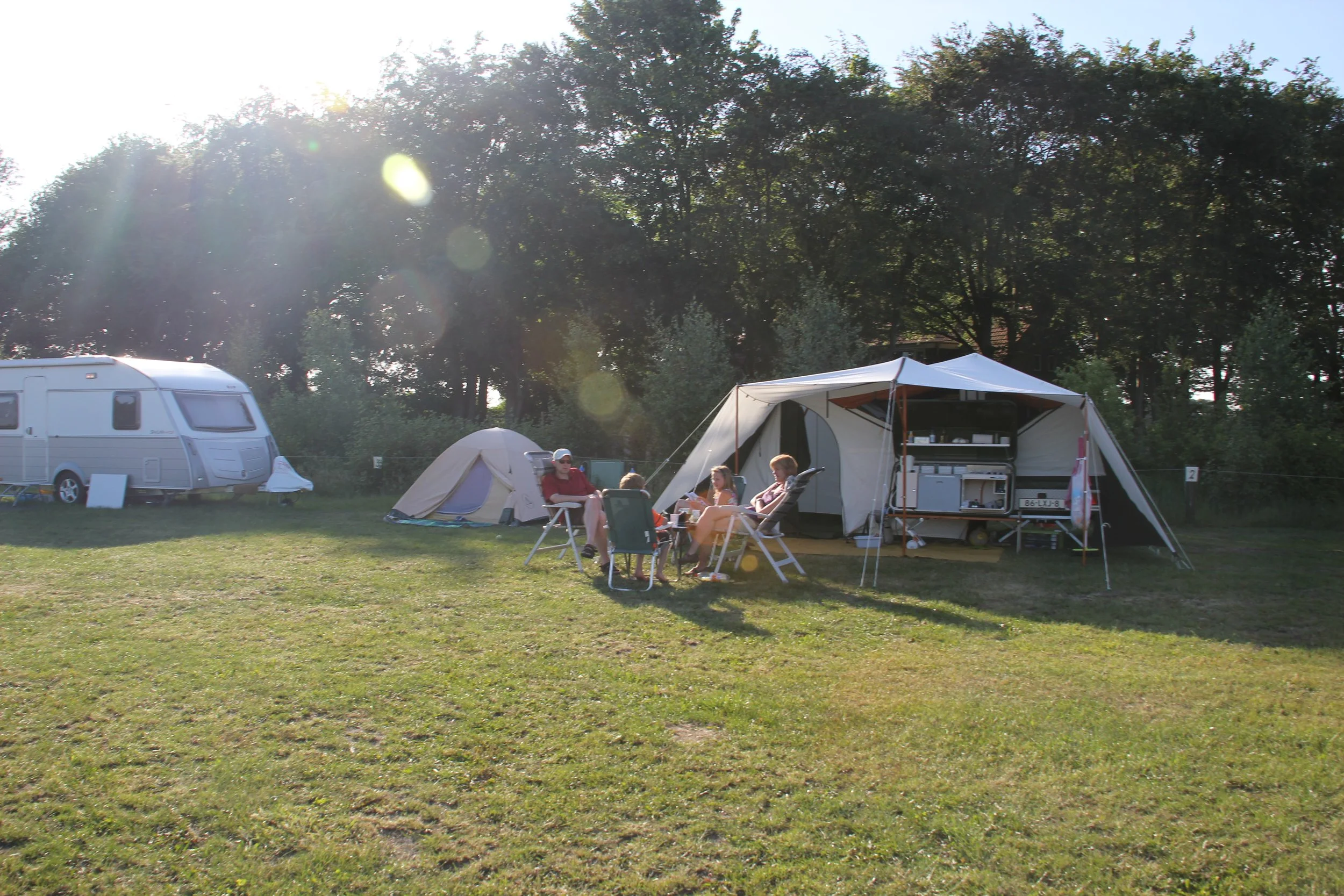 Mensen zitten samen op een camping bij een grote tent, met een camper op de achtergrond en bomen eromheen, bij zonsondergang.