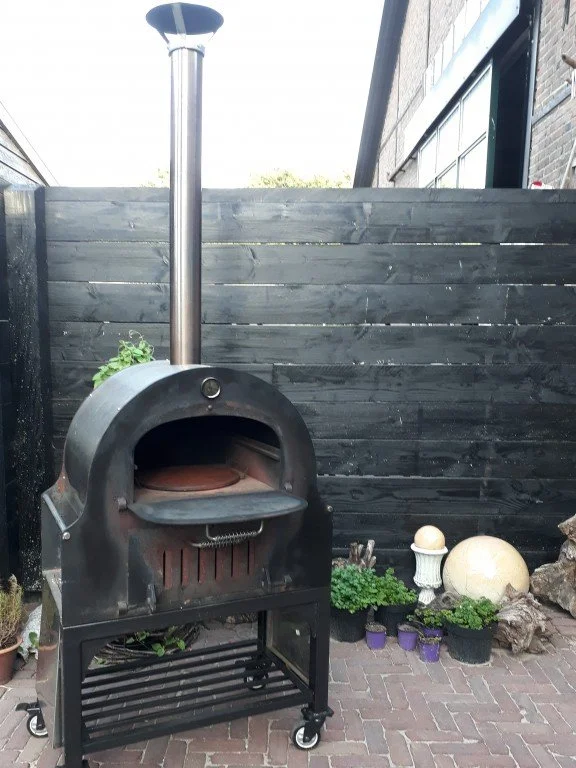 Een buitenkeuken met een pizzaoven op wielen, geplaatst op een stenen patio, omgeven door planten en decoratieve objecten, met een houten schutting op de achtergrond.