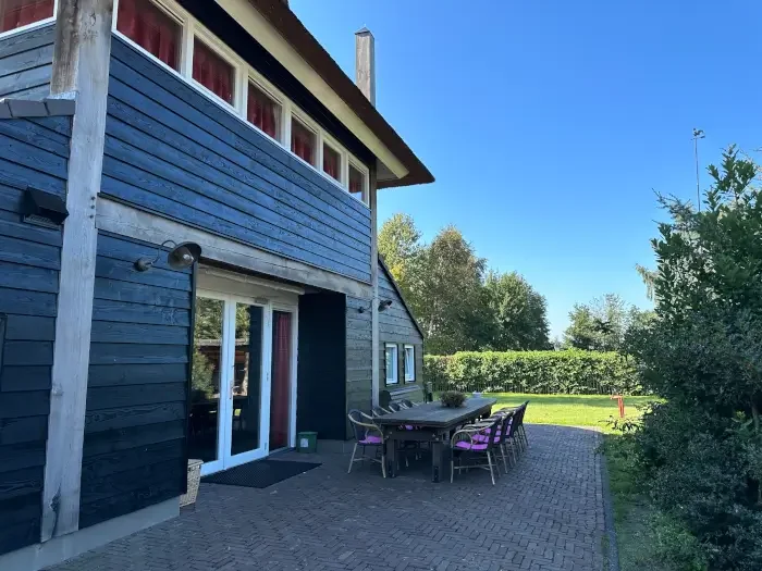 Een huis met een houten gevel in donkerblauwe kleur, een tuin met terras en tuinmeubilair onder een heldere blauwe hemel.