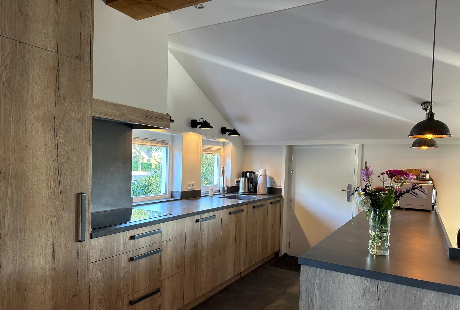 Een moderne keuken met houten kasten, twee ramen, een werkblad met koffiezetapparaat en bloemstuk, en een plafond met zwarte lampen.