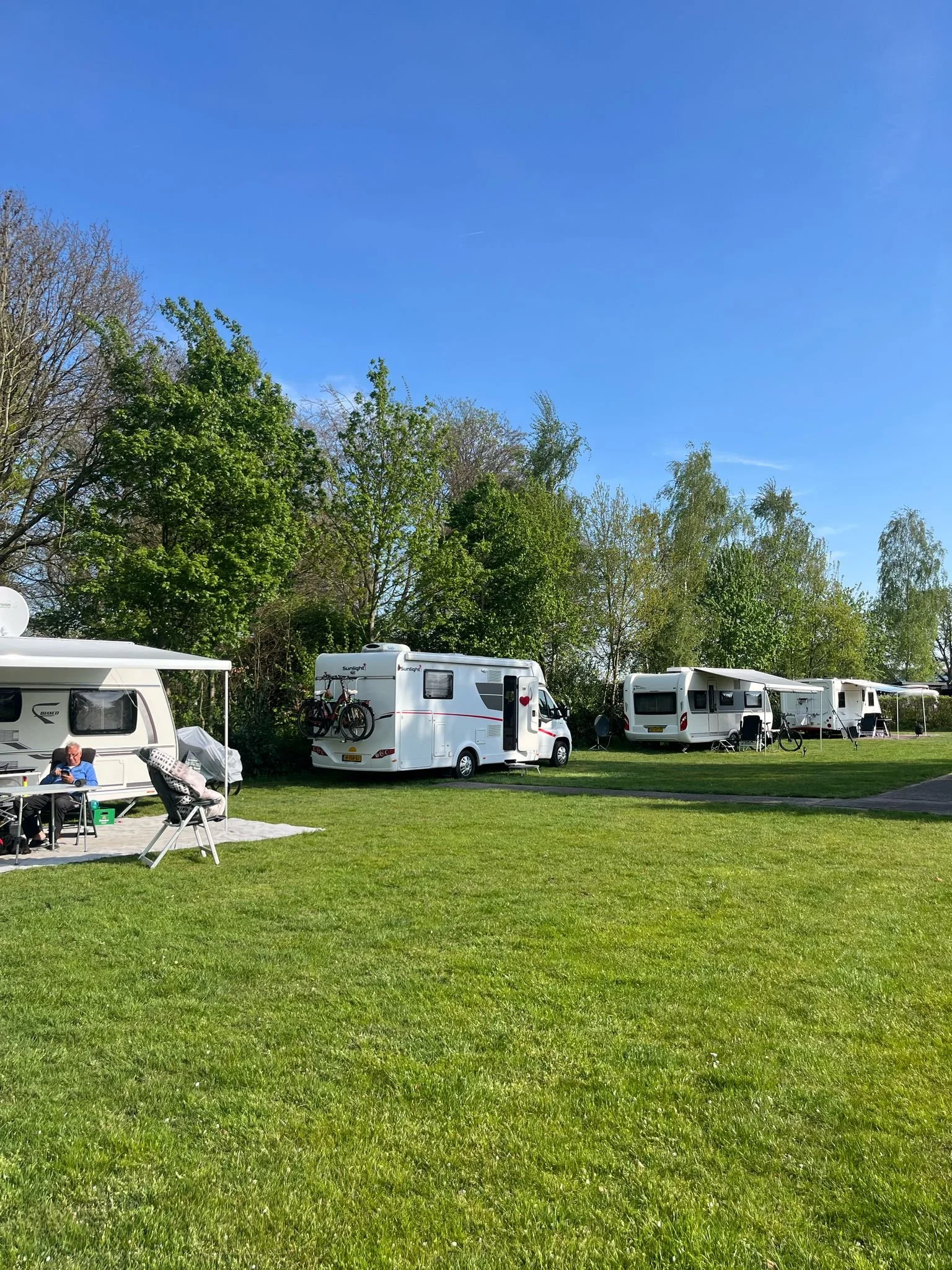 Campingsite met caravans en campers onder een blauwe hemel met groene bomen op de achtergrond.