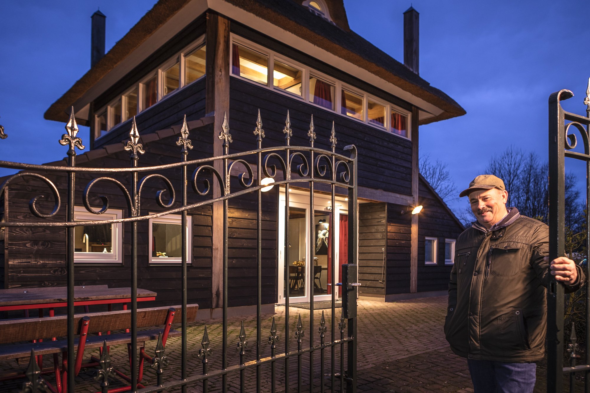 Man staat buiten bij een woning 's avonds, achter een smeedijzeren poort, de woning is een twee verdieping huis met houten gevelbekleding en grote ramen, het is donker buiten en het huis is verlicht.