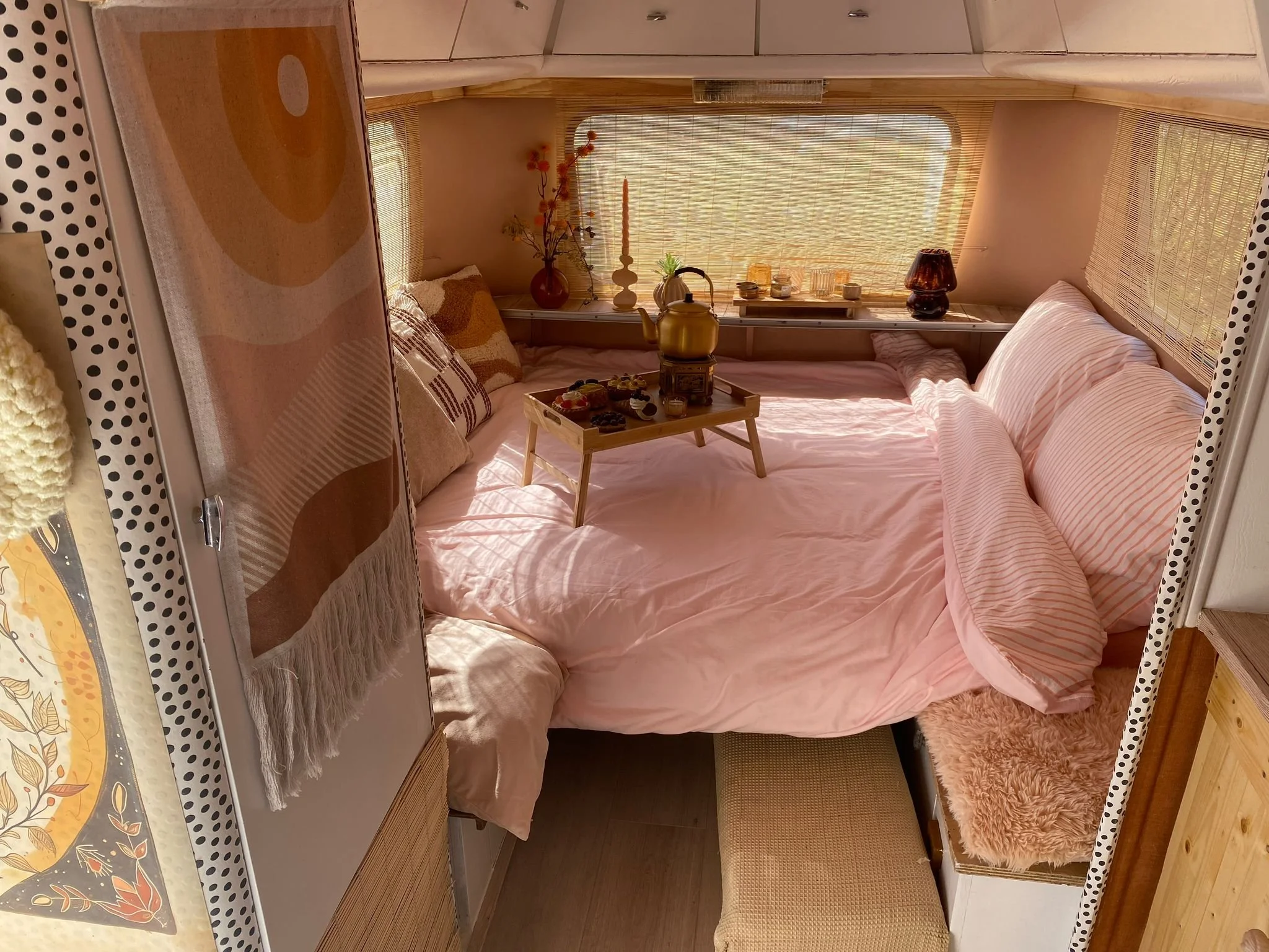 Knus kleine slaapkamer met een bed in roze en witte strepen, versierd met kussens en een tapijt, met een achtergrond van een raam met houten jaloezieën en decoratieve accessoires zoals een vaas met bloemen en kaarsen.