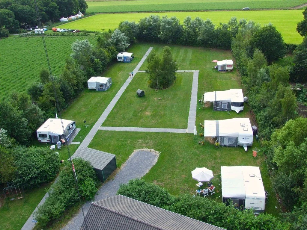 Vijf caravans op een grasveld, omringd door bomen en struiken, met paden en een terras met een parasol en zitjes.