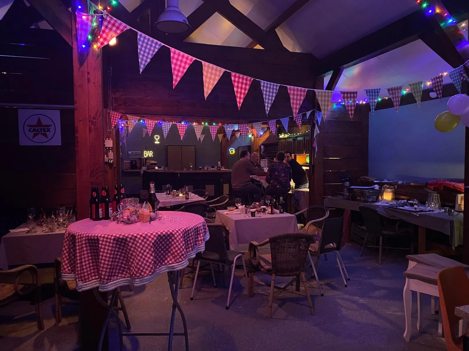 Binnenkant van een gezellig feestcafé met feestversiering, zoals kleurrijke vlaggetjes en lichtjes, tafels met tafelkleedjes, en een bar in de achtergrond waar mensen samen zitten.