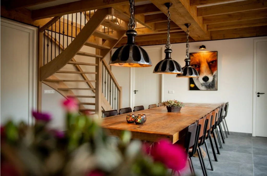Een eetkamer met een lange houten tafel, zwart gekleurde stoelen, hangende zwarte lampen, een schilderij van een vos aan de muur, en een houten wenteltrap in de achtergrond.