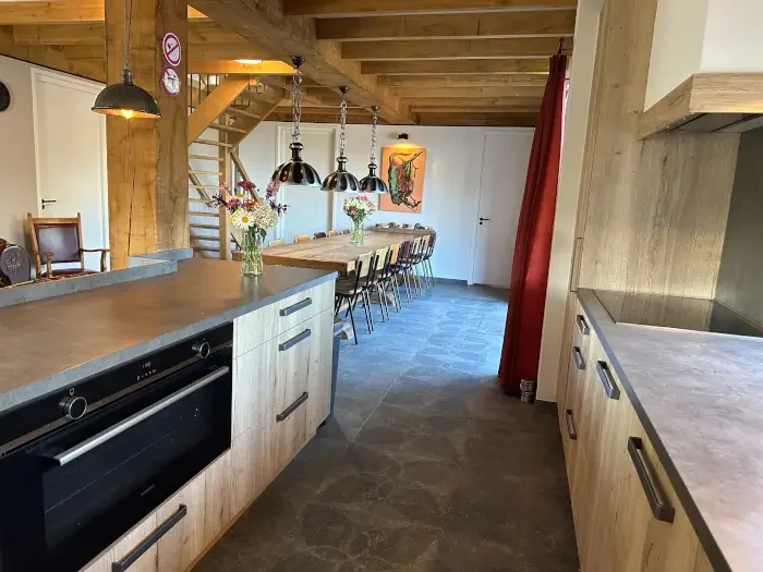 Interieur van een moderne keuken met een eettafel en lange bank, houten plafond en stalen hanglampen, bloemen in vazen, kleurrijk kunstwerk op de muur, trap naar boven, houten kasten en een kookplaat.