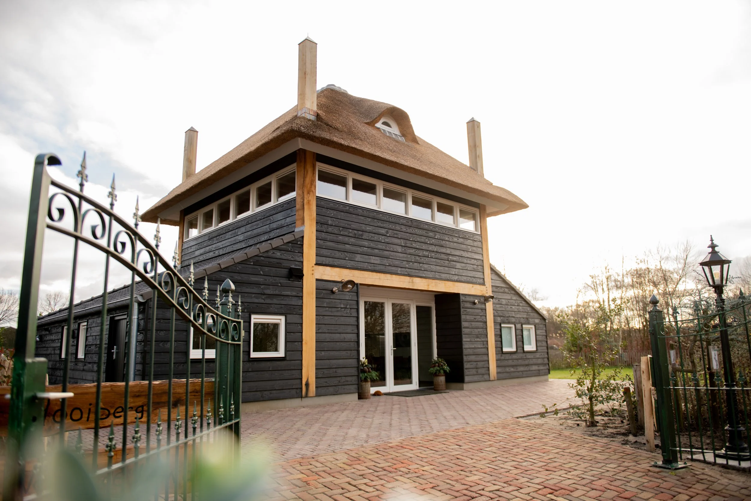 Een uniek twee verdiepingen huis met zwarte houten gevel en rieten dak, omgeven door een oprijlaan en tuin, met een hek en lantaarnpaal voor de ingang.