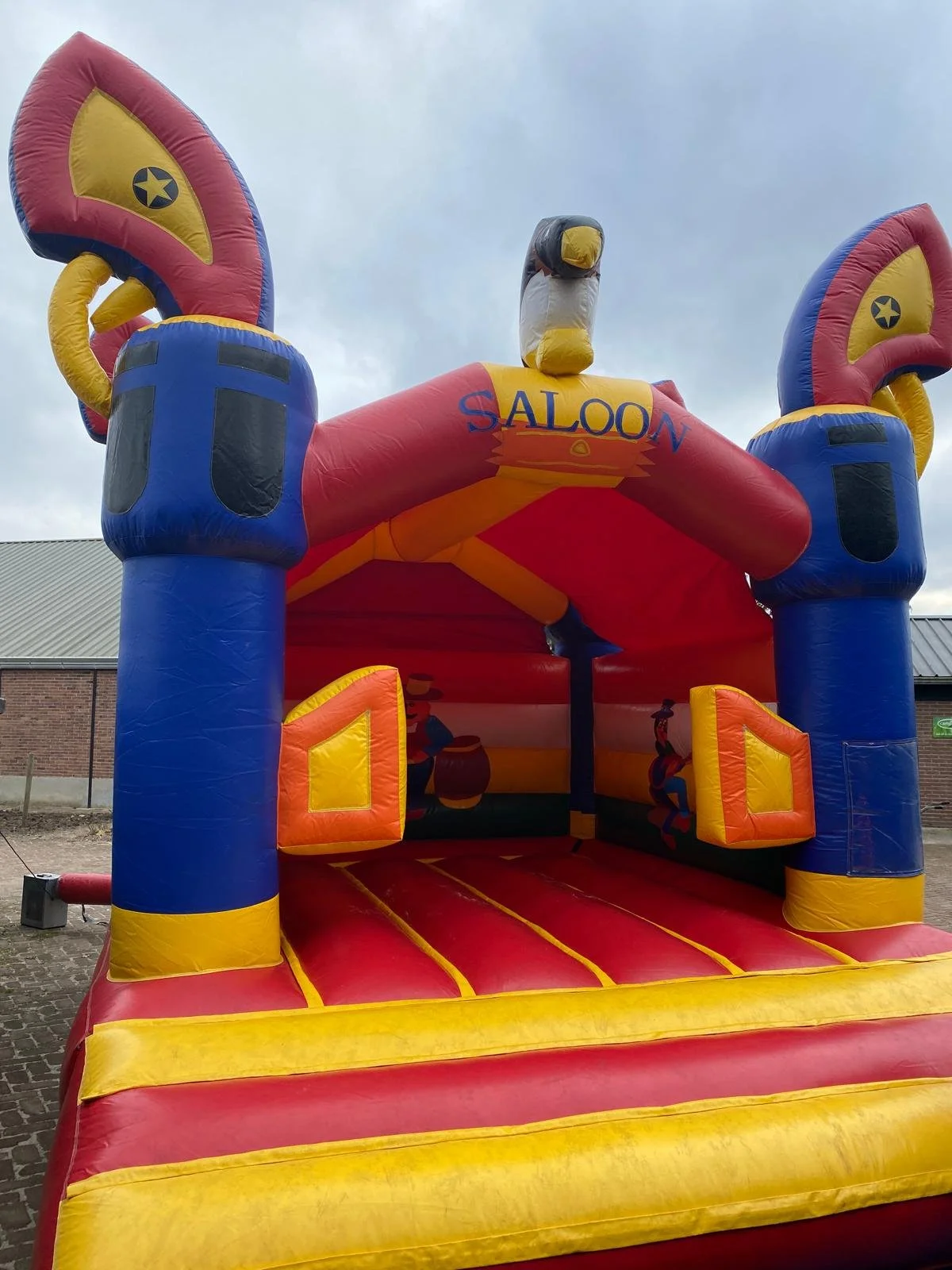 Grote rode, gele, blauwe en zwart springkussen met een western saloon thema, met tekeningen van cowboys en cowgirls en een grote buizerd op de top.